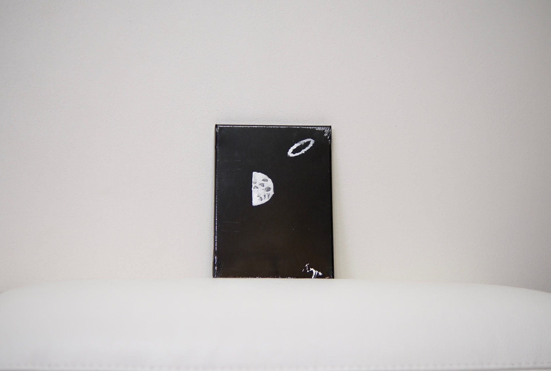 The small black canvas standing on a white cushioned surface against a pale wall, dwarfed by the empty space around it.