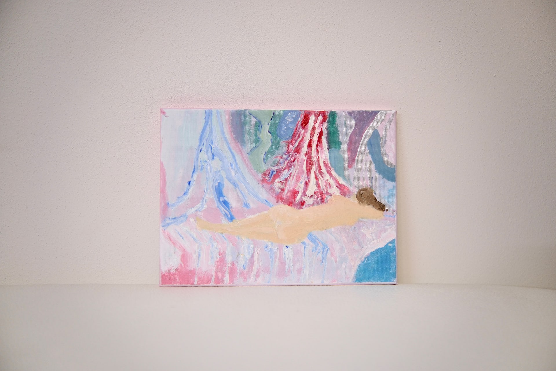 The unframed canvas leaning against a pale wall, its reclining pale figure set against streaming pink and blue.